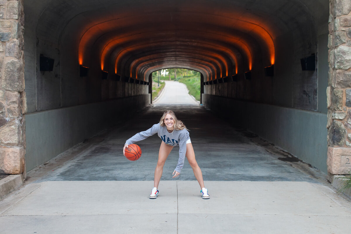 A person wearing a gray sweatshirt and white shoes is dribbling a basketball in a tunnel with glowing orange lights. The tunnel leads to a green, sunlit area in the distance.