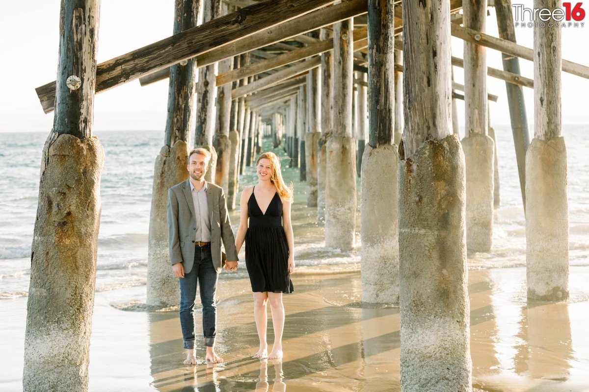 Engaged couple hold hands while standing under the Newport Beach Pier with their feet in the water