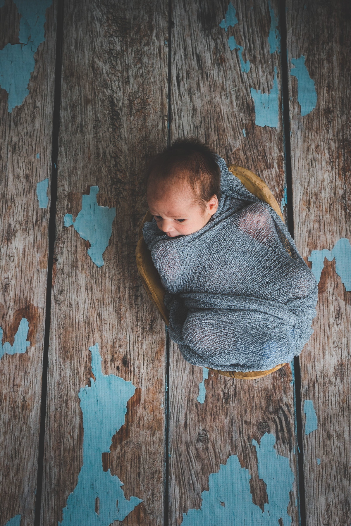 Newborn photography of baby wrapped in cheesecloth blanket in a bowl in a wooden floor.