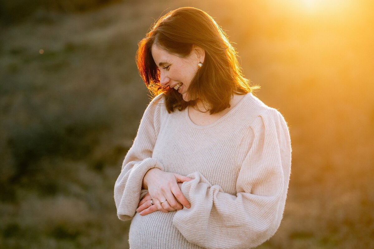 Zwangere vrouw poseert lachend terwijl ze naar beneden kijkt in de golden hour