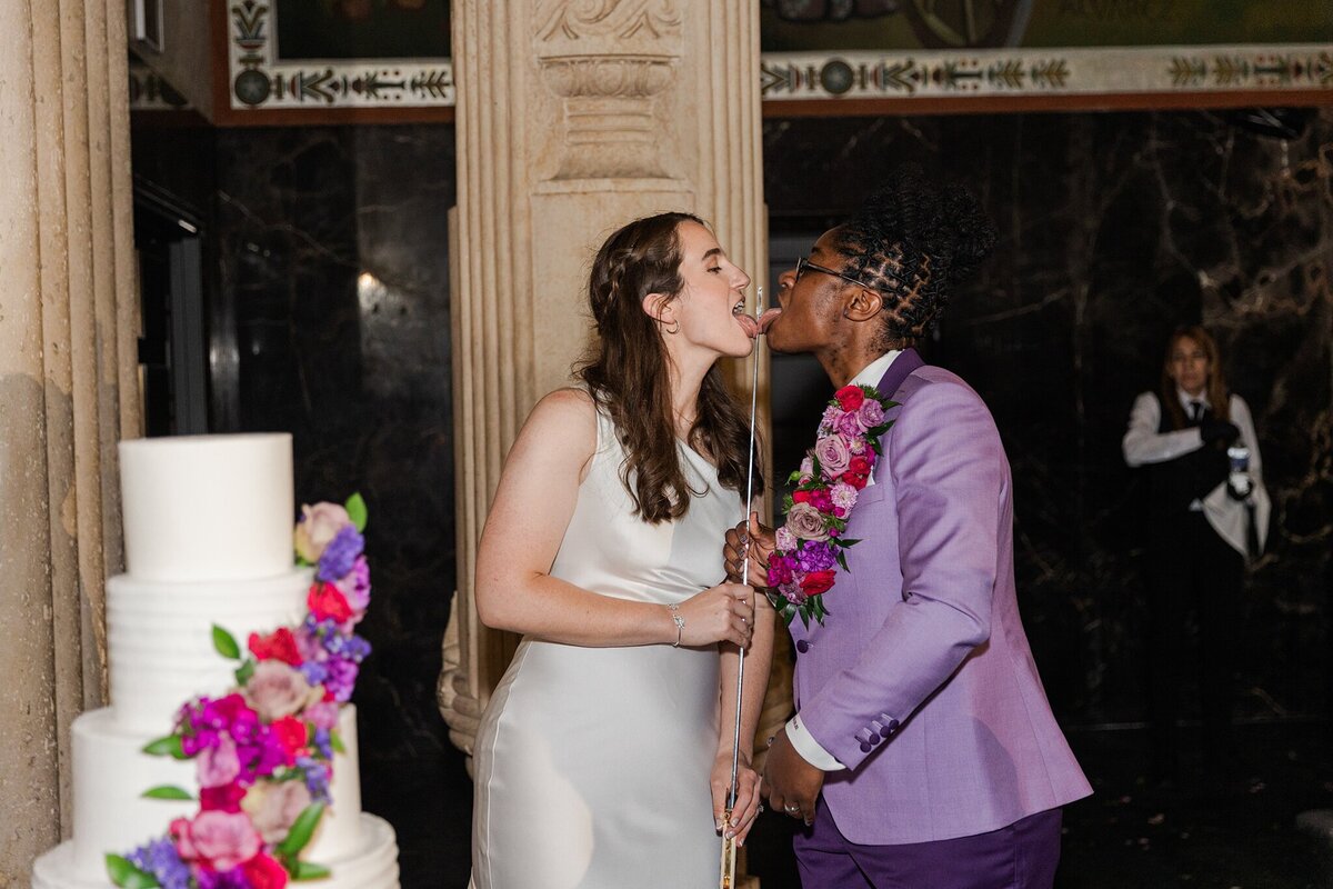 Two brides licking a sword that they used to cut their wedding cake during their wedding reception at the Hall of State in Fair Park in Dallas, Texas. The bride on the left is wearing a sleeveless, elegant, white dress, and the bride on the right is wearing a purple suit with a very large boutonniere.