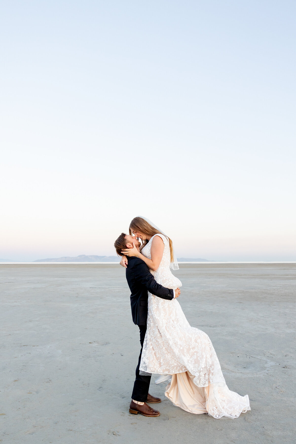 Groom lifts the bride into the air as they embrace, surrounded by an open desert-like landscape.