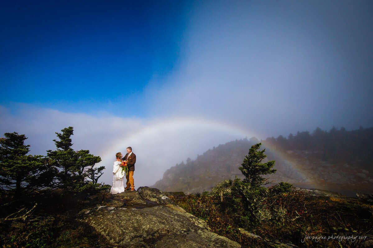 A wedding couple standing on a rock together with a rainbow behind them.