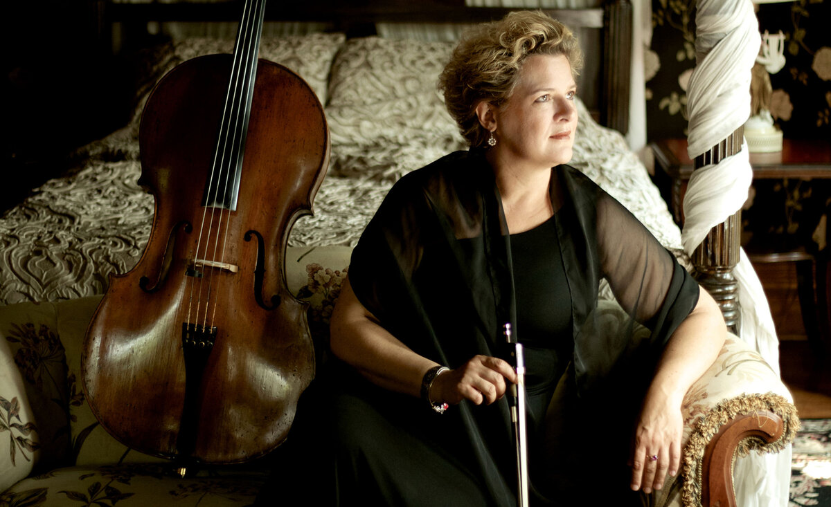 Classical musician portrait Margaret Maria wearing black dress sitting against white sofa beside cello