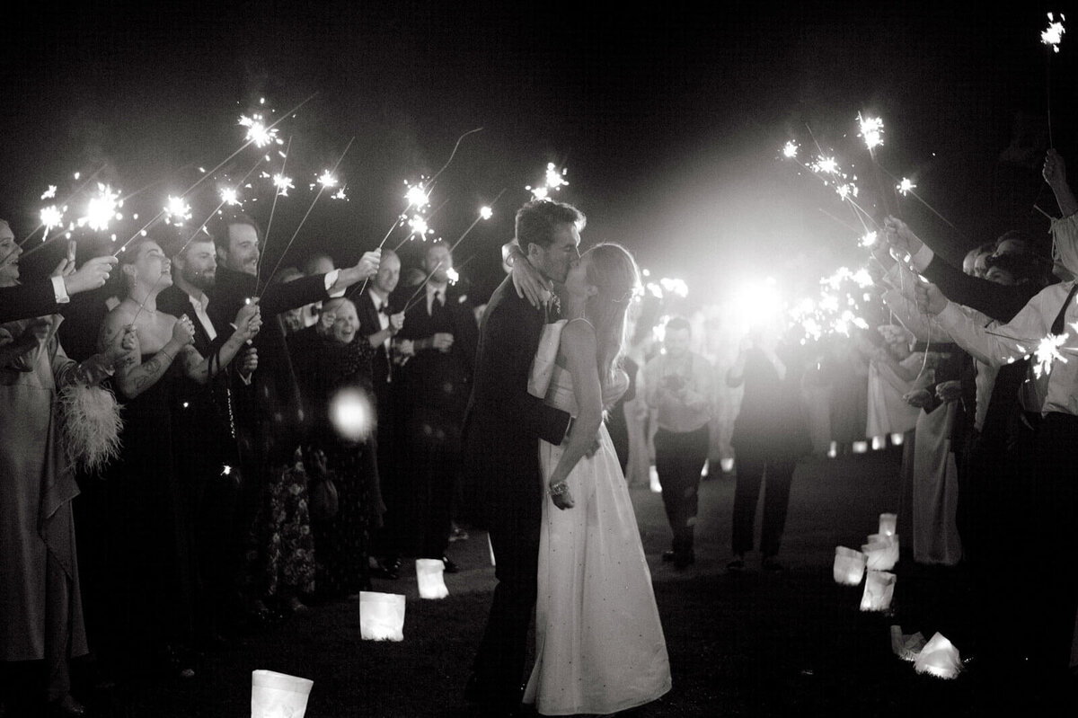The bride and the groom kissed outside of the tent wedding venue as guests watched, holding sparklers, at The Ausable Club, NY.