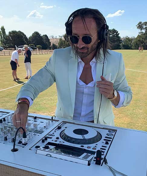 Male DJ in a pastel suit playing music at an outdoor polo field during a Melbourne Cup event