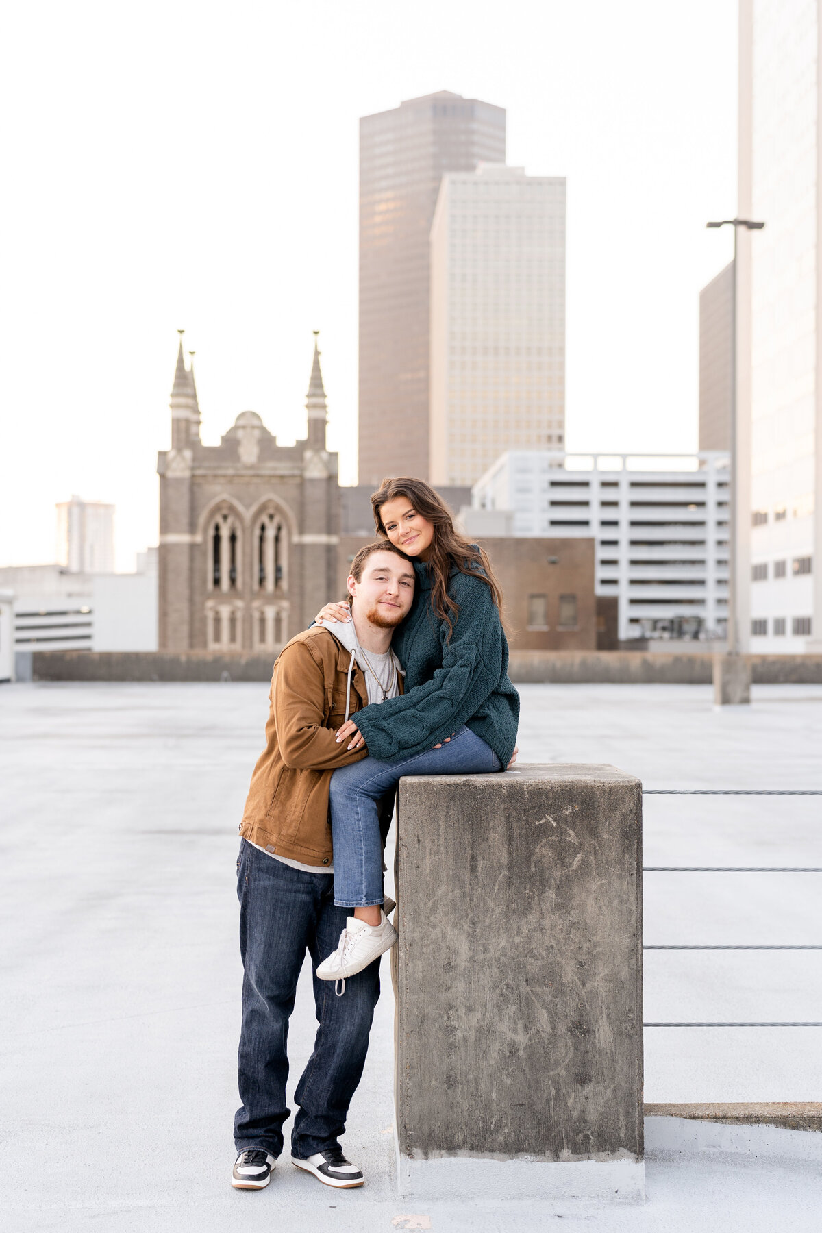 Future bride and groom smiling at camera while girl is sitting on concrete podium on top of a rooftop parking garage with Downtown Houston in the background