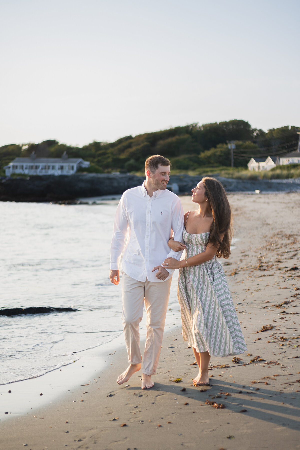 Collins Beach Newport RI | Kelsey Sheehan Photography Timeless Rhode Island Weddings | A couple walks barefoot along a sandy beach at sunset, holding hands and smiling at each other. They are dressed in light, casual summer clothing.
