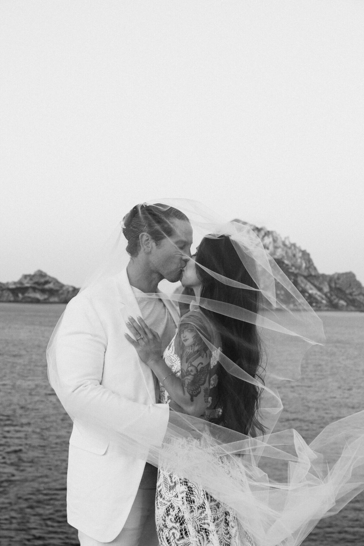 Couple in wedding clothes kissing under a veil with Es Vedra in the background