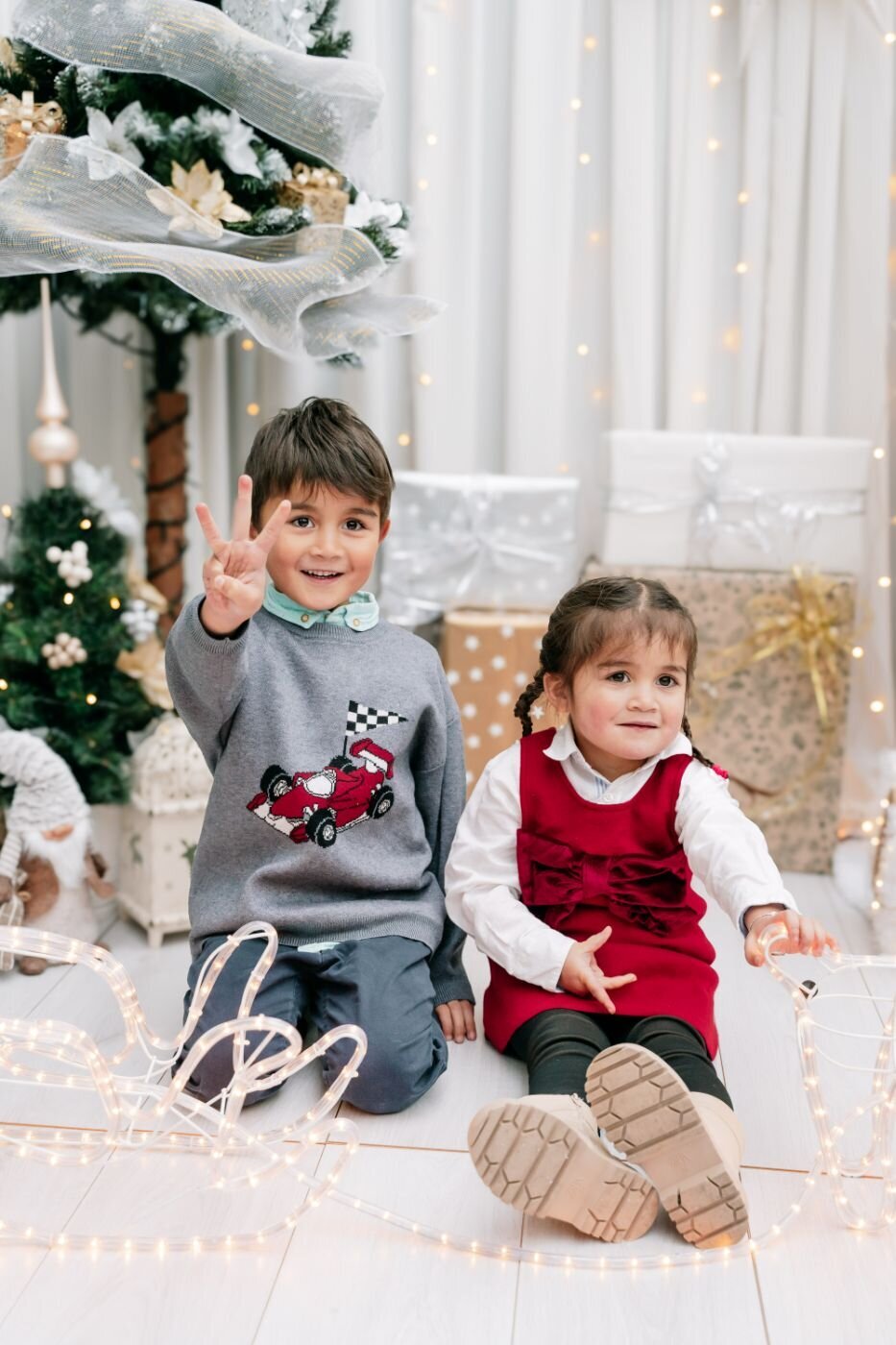 Two children at Christmas – Young boy flashing a playful hand sign and little girl in a red dress sitting by a Christmas tree surrounded by twinkling lights and wrapped presents.