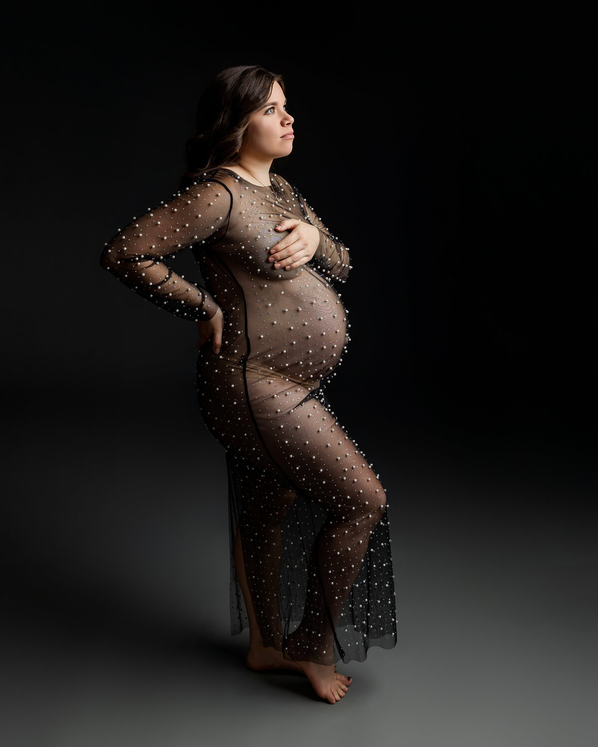 A stunning mother-to-be wearing a black sheer dress adorned with delicate pearls, captured in a side view against a soft, dark backdrop. Captured by Inland Empire maternity photographer Kaitlyn Dawn Photography.