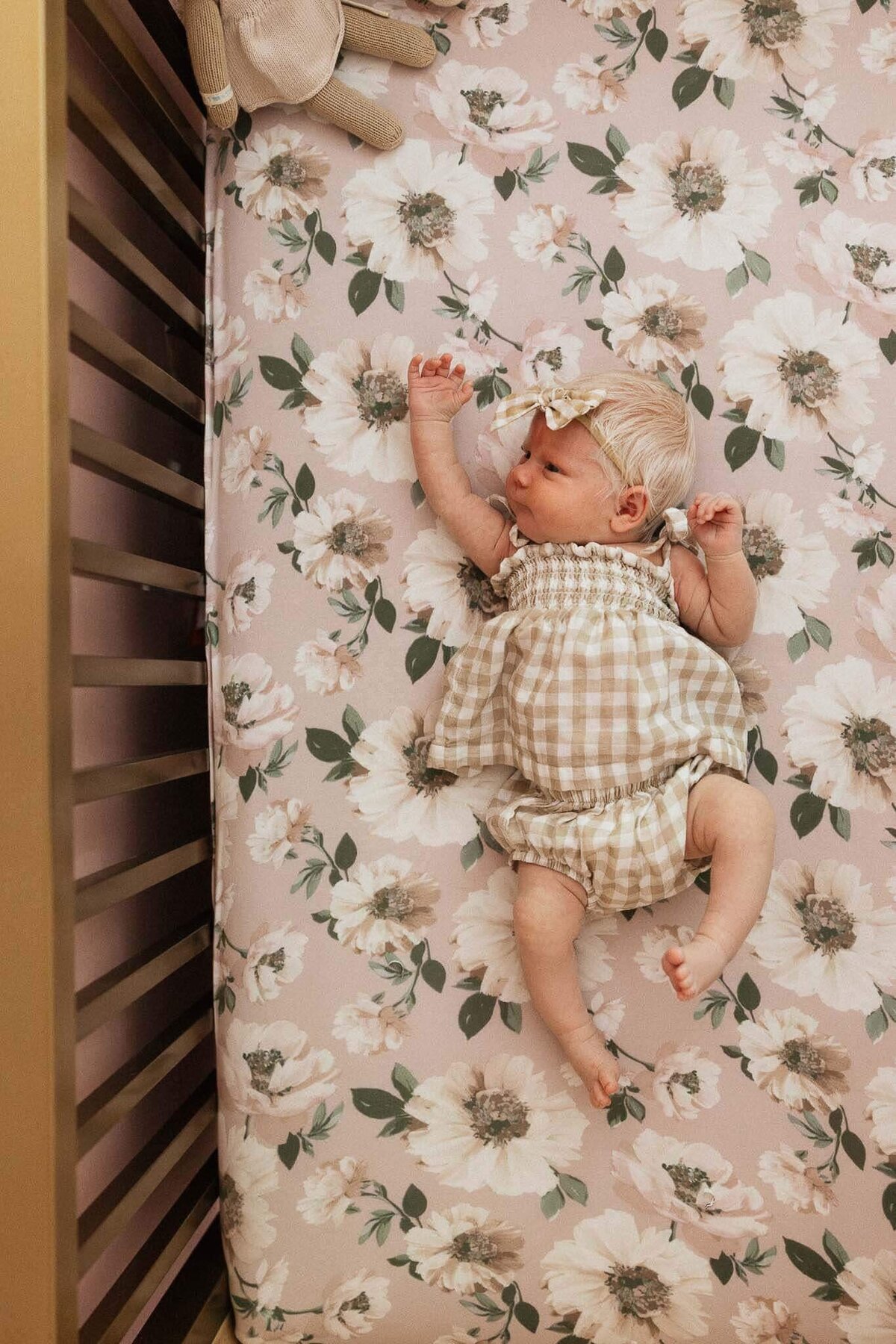 A newborn dressed in a gingham outfit lies in a floral-themed crib, showcasing a cozy and vibrant nursery style during Cleveland in-home newborn photography session.