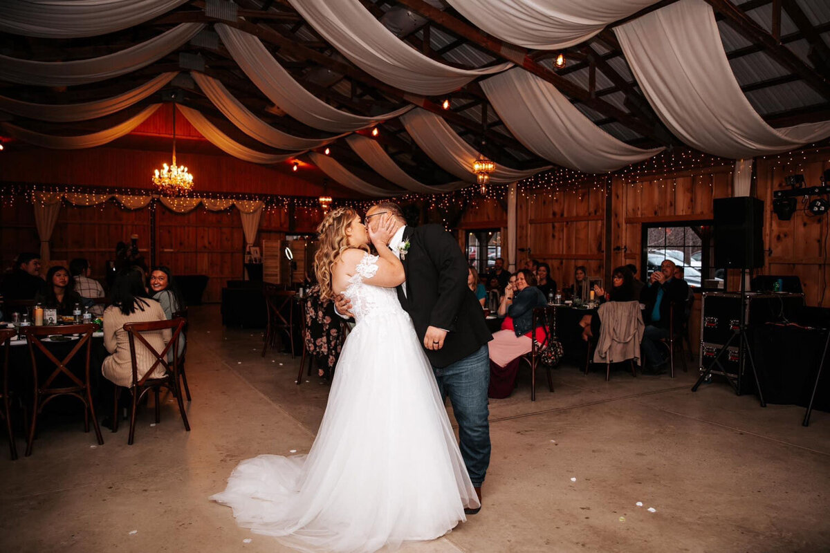 Bride and groom sharing a kiss on the dance floor in a warmly lit barn with guests seated