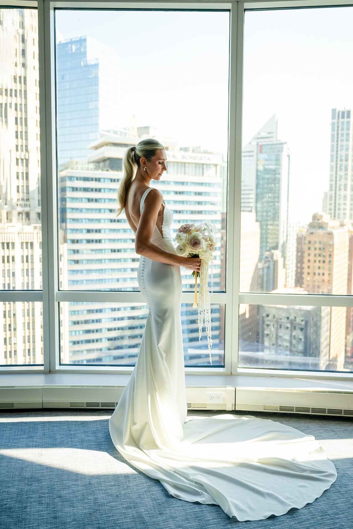 Wife in white wedding dress holding flowers looking outside at buildings of Chicago from Trump Hotel