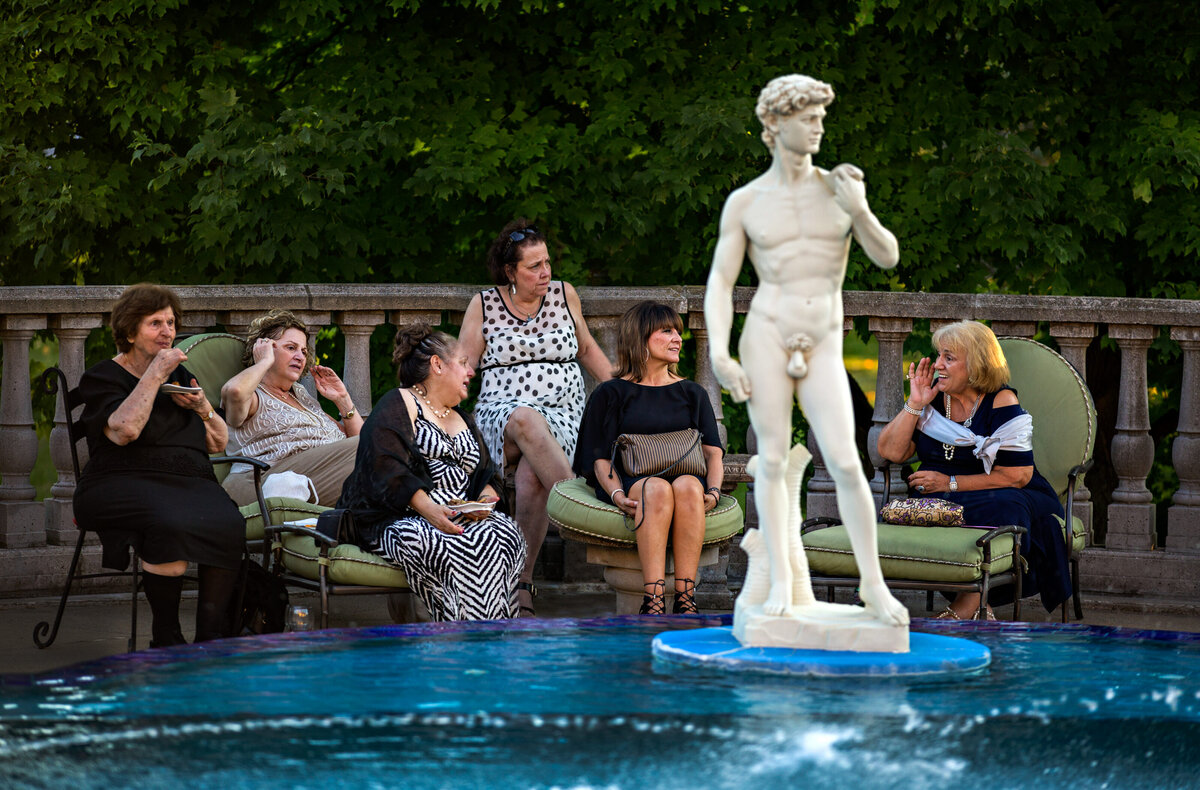 Ladies eat during cocktail at a swimming pool with a Greek statue in front of them.