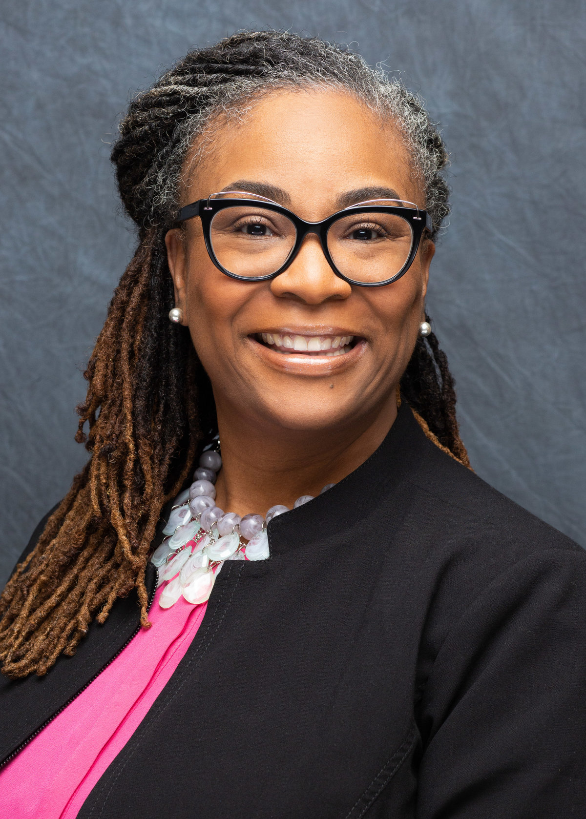 Vibrant headshot of a joyful black woman with elegant dreadlocks, stylish glasses, and a pink blouse, professionally photographed in Cincinnati.