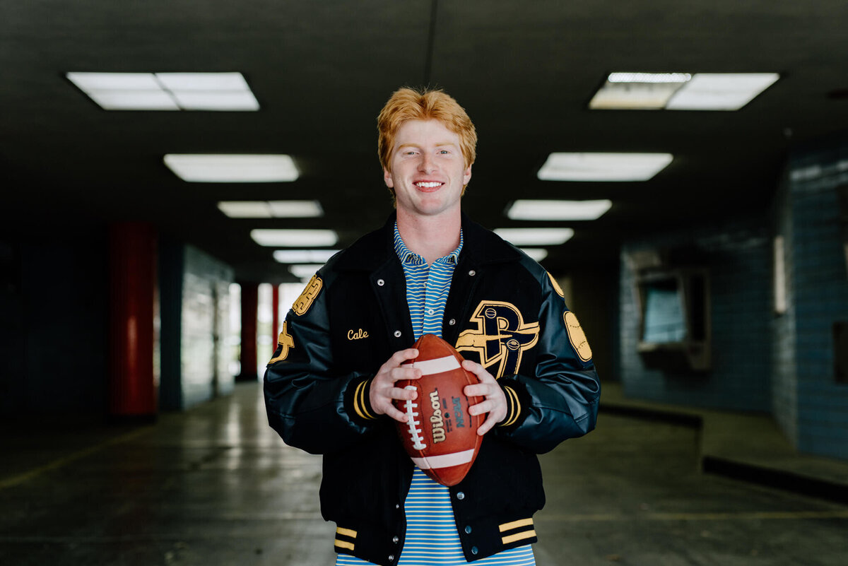 senior portrait of Pine Tree High School football player wearing letterman jacket and holding football in Longview, TX parking garage