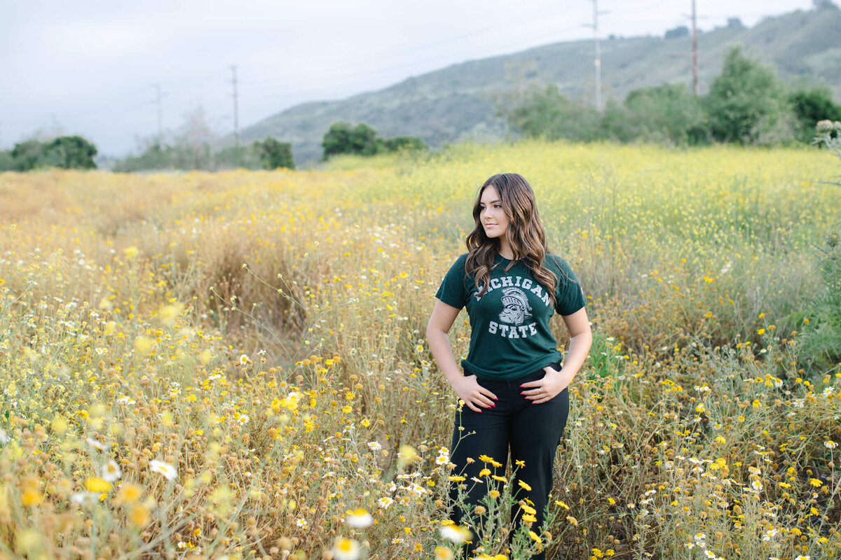 A woman with long, brown hair wearing a green Michigan State shirt and black jeans, with her thumbs in her belt loops, looking off to her right side in a field of yellow flowers and green grass in Northwest Open Space in San Juan Capistrano.