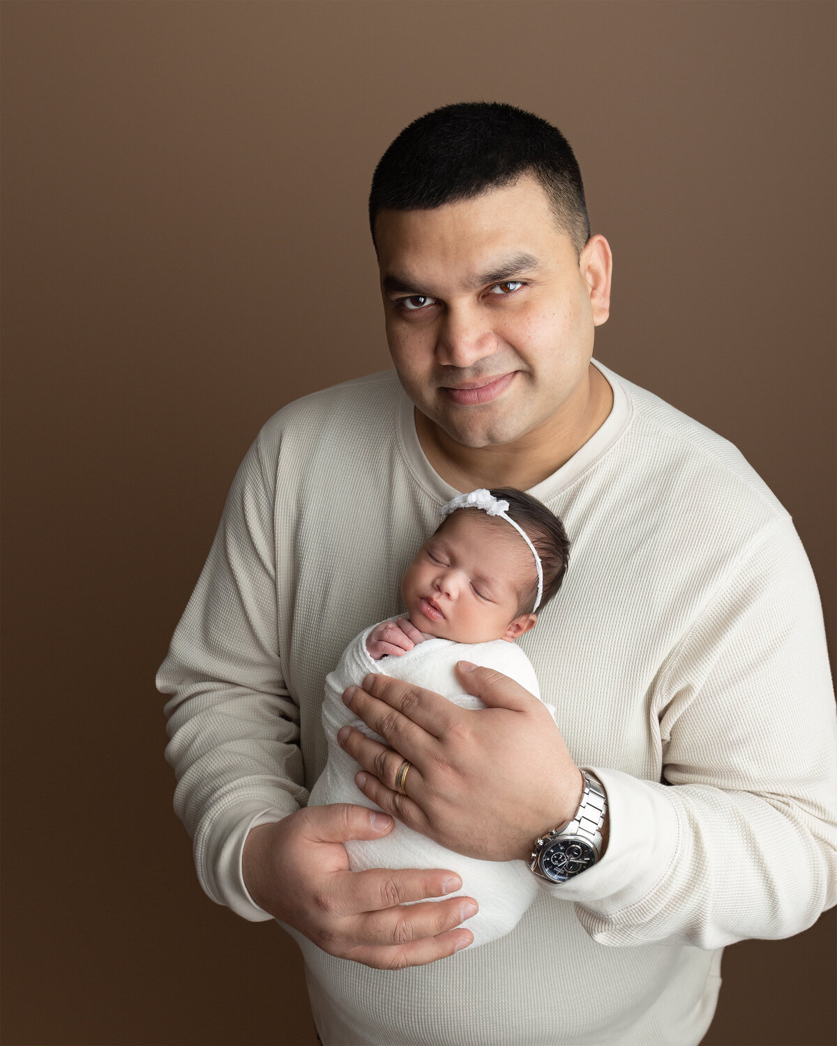 A tender studio portrait of a father cradling his newborn daughter in front of a warm brown backdrop. Photographed in my studio in Paris, Ontario.