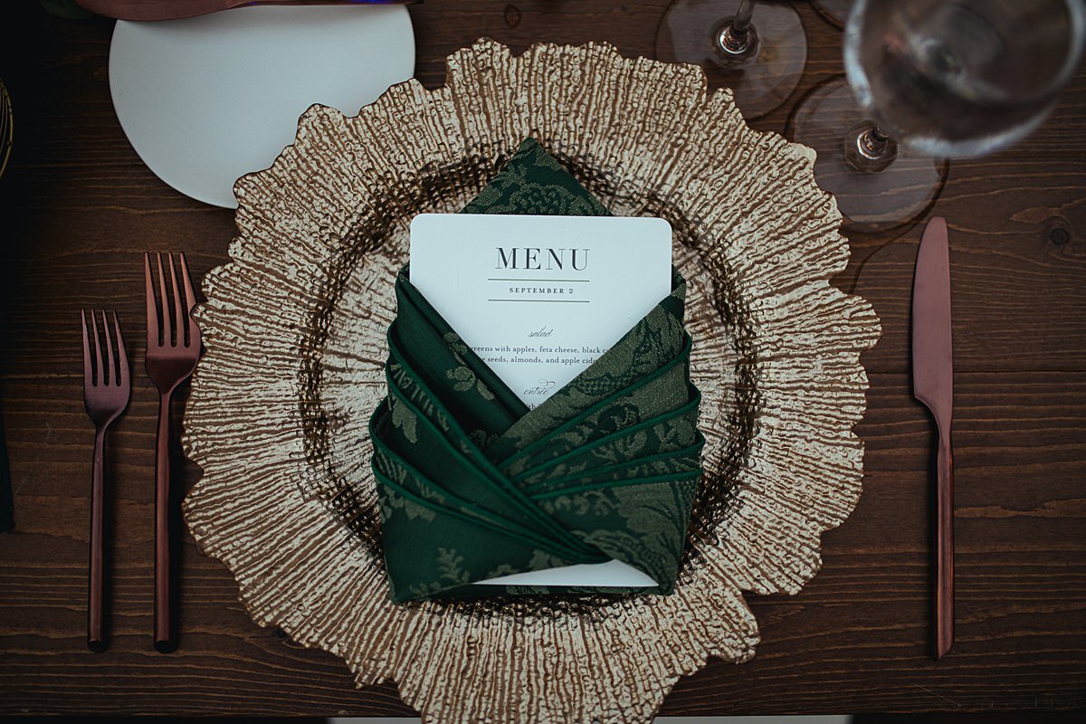 An ivory menu card wrapped in a forest green brocade napkins on a gold glass charger set on a dark wood table with a bread plate, copper flatware and a long stemmed wine glass place setting