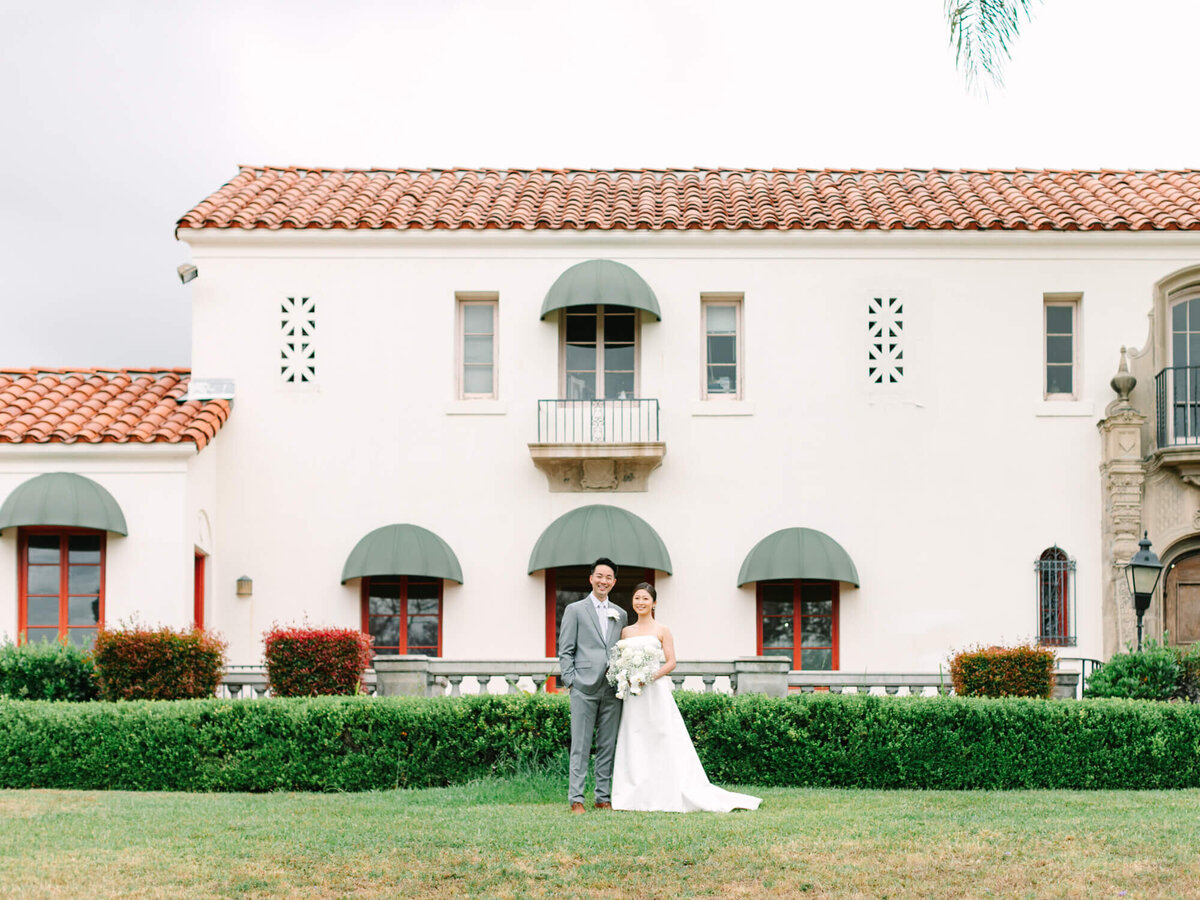 Bride and groom stand smiling in front of an elegant white building with red roof tiles and green awnings, lush greenery in foreground, conveying joy.