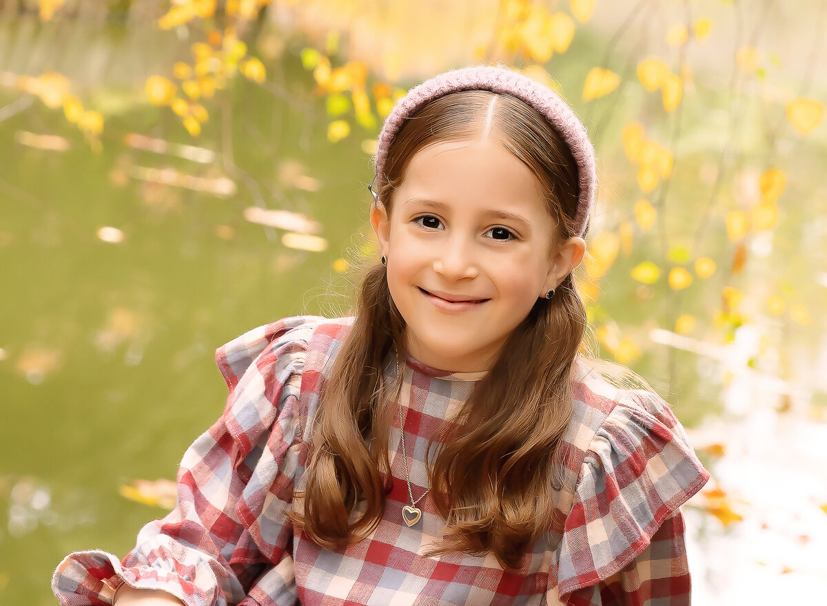 Little girl in a red plaid dress is sitting in front of a pond for a fall family photoshoot. Captured by Brooklyn family photographer Chaya Bornstein