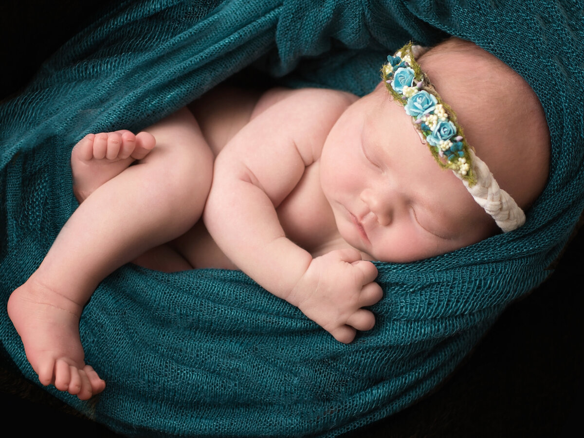 Naked baby girl with rolls cuddled up in a teal swaddle wrap.  She has a headband with small teal flowers.  Her leg is hanging out and her arm is clutcing the wrap.  She is asleep.