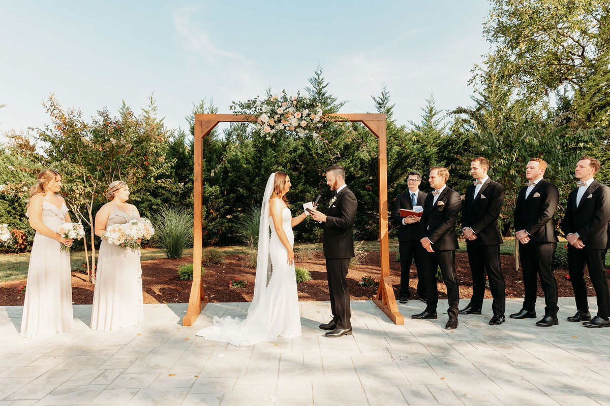 A bride and groom stand under a wooden floral arch at their private event venue in Northern Virginia, exchanging vows. The bride is in a white dress, and the groom in a black suit. Bridesmaids in light dresses and groomsmen in suits stand nearby on this sunny day, surrounded by lush greenery.