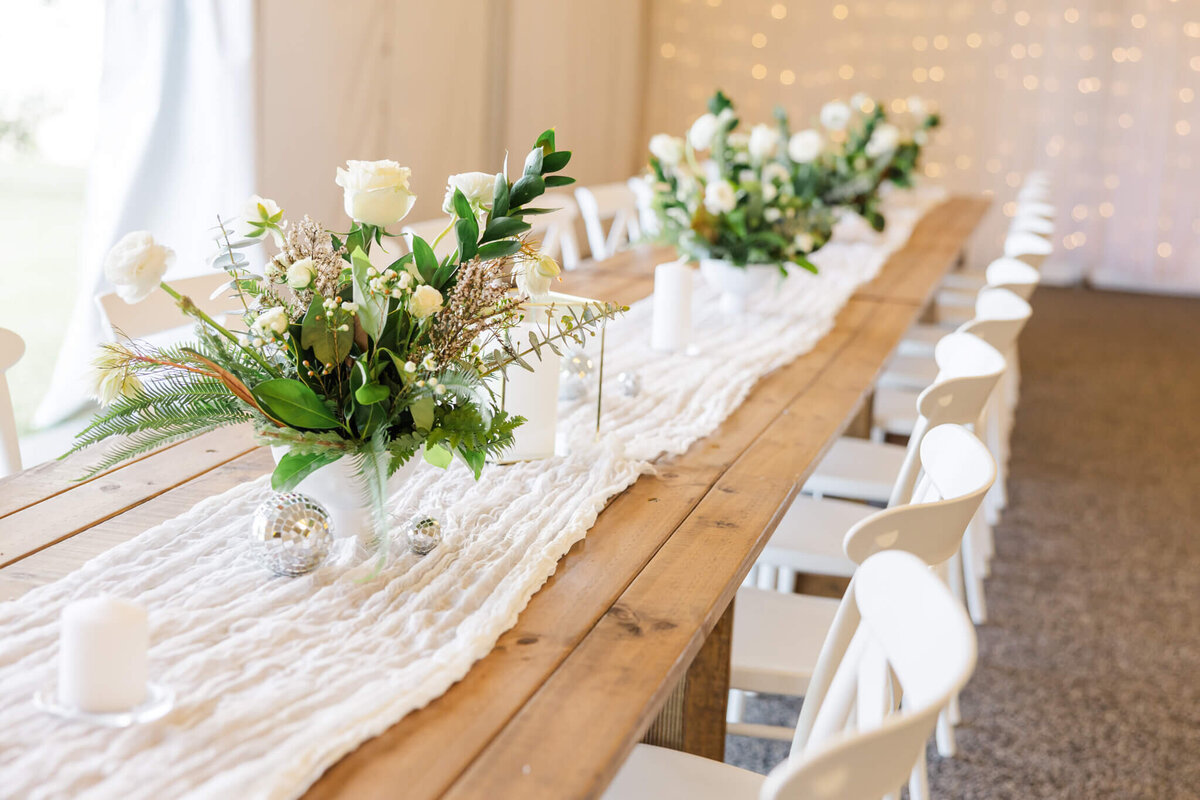 A long wooden table set up for a wedding reception with a white cheese cloth runner and green and cream floral arrangements as centrepieces.