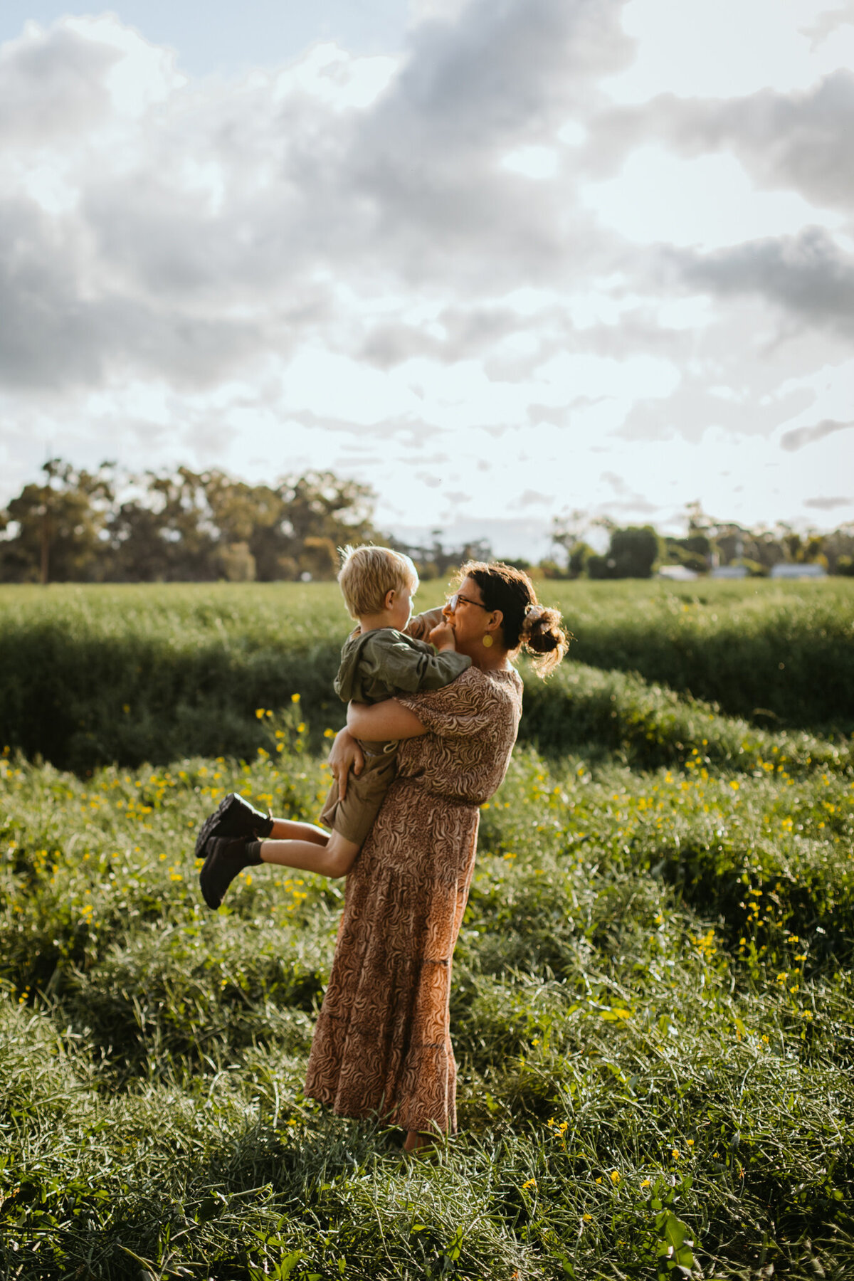 Son wraps his arms around his mother’s neck as she lifts him in a sunlit, flower-filled field.