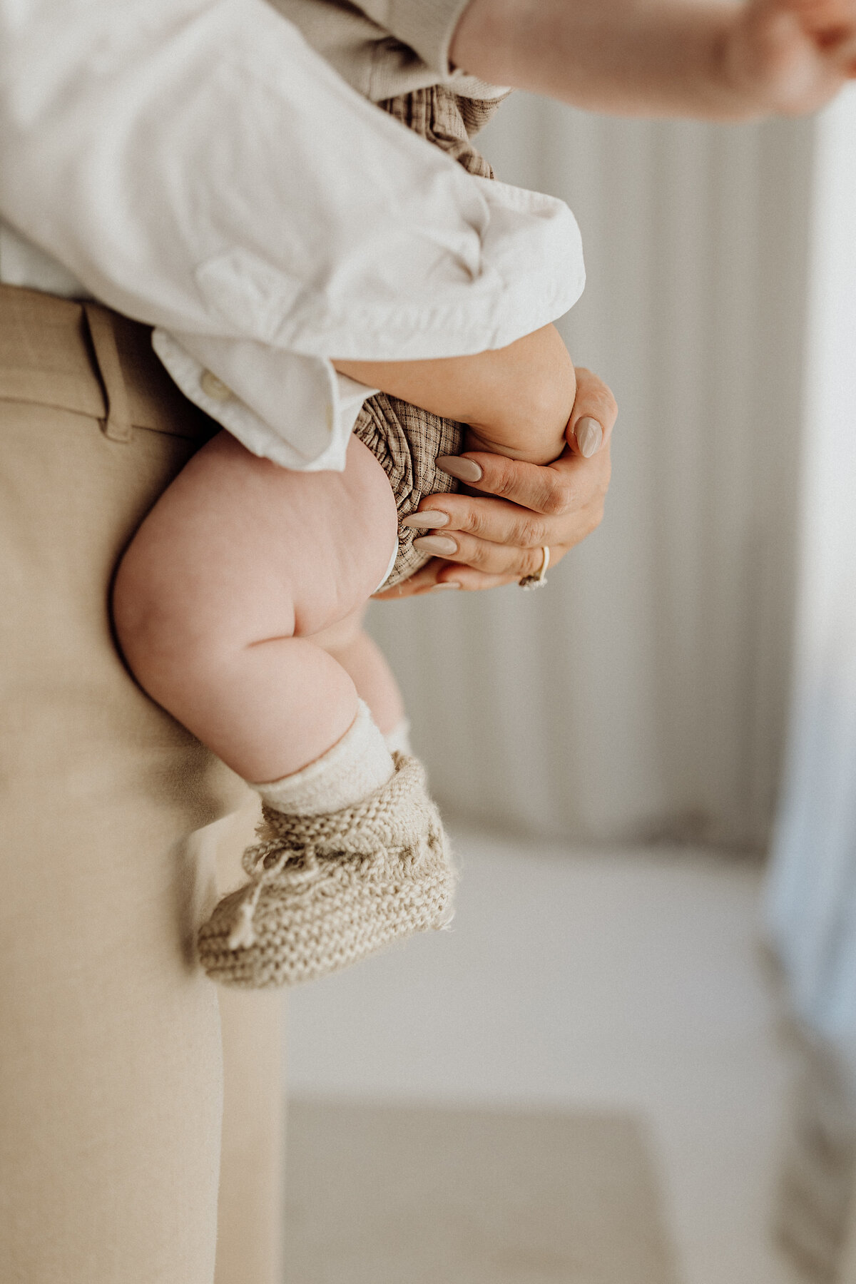 A mother holding her baby wearing knitted booties, styled in cream and white clothing and standing in lucys newborn studio in christchurch