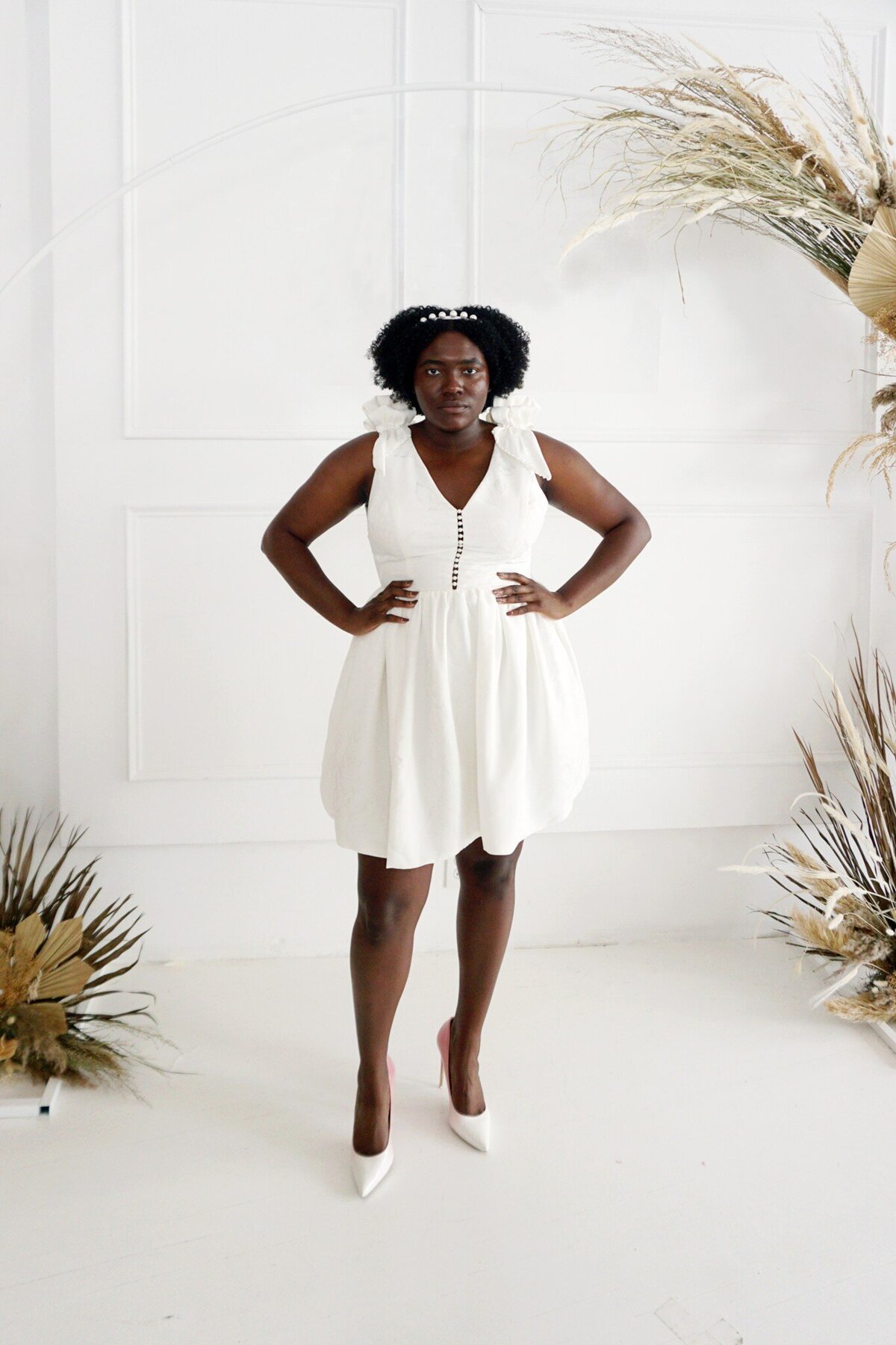 Black model wearing a size-inclusive little white wedding dress. The style features pearl embellishments and shoulder ruffles.