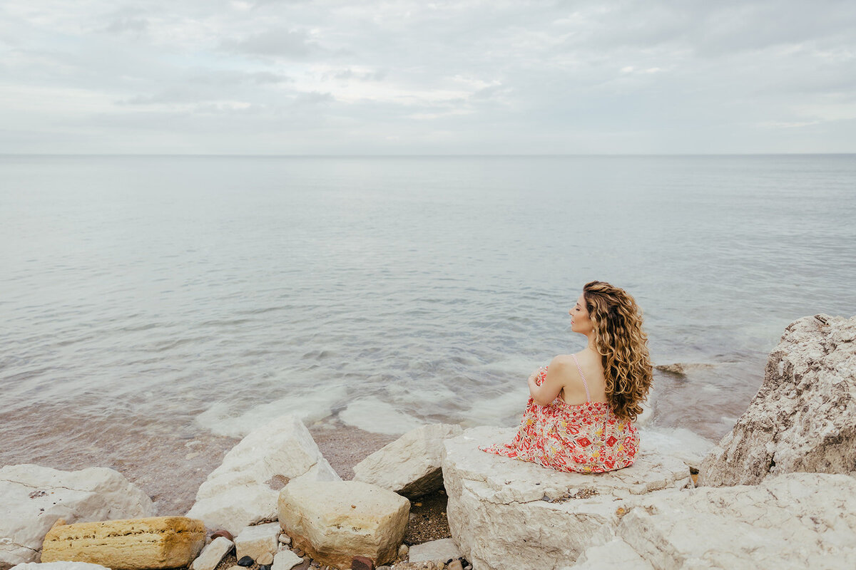 Woman in red dress is sitting on rocks looking out over Lake Michigan