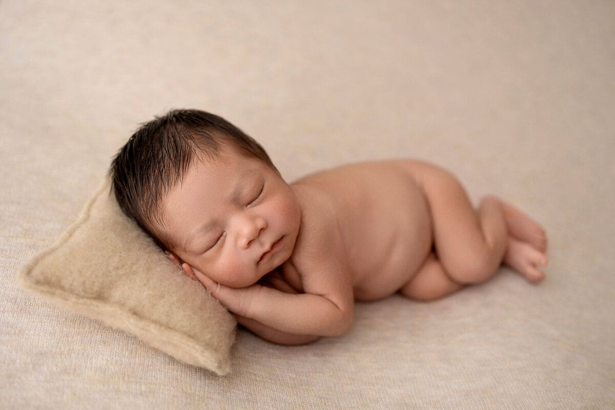 Newborn baby peacefully sleeping on a beige blanket, resting head on a small beige pillow.