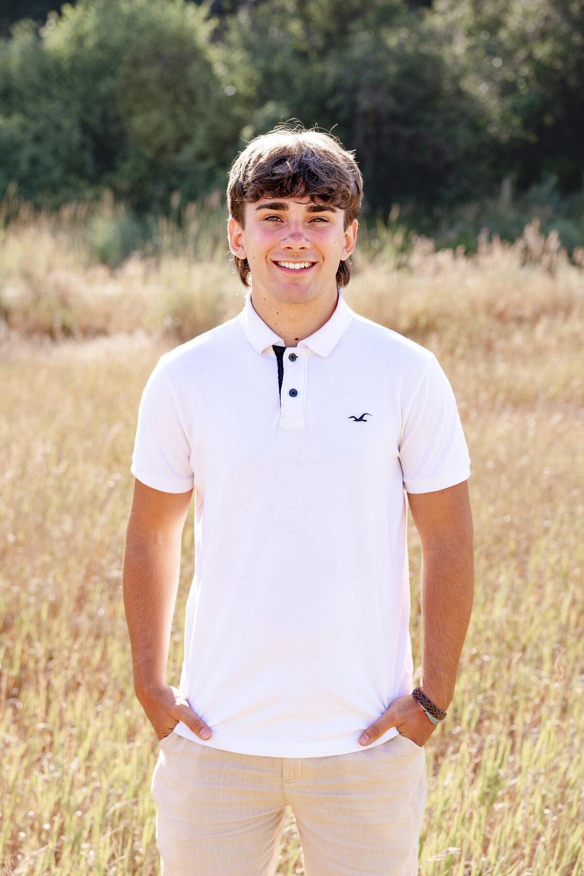 A teenage boy in a white polo shirt and beige pants smiles at the camera, standing in an open meadow with warm sunlight filtering through the trees.