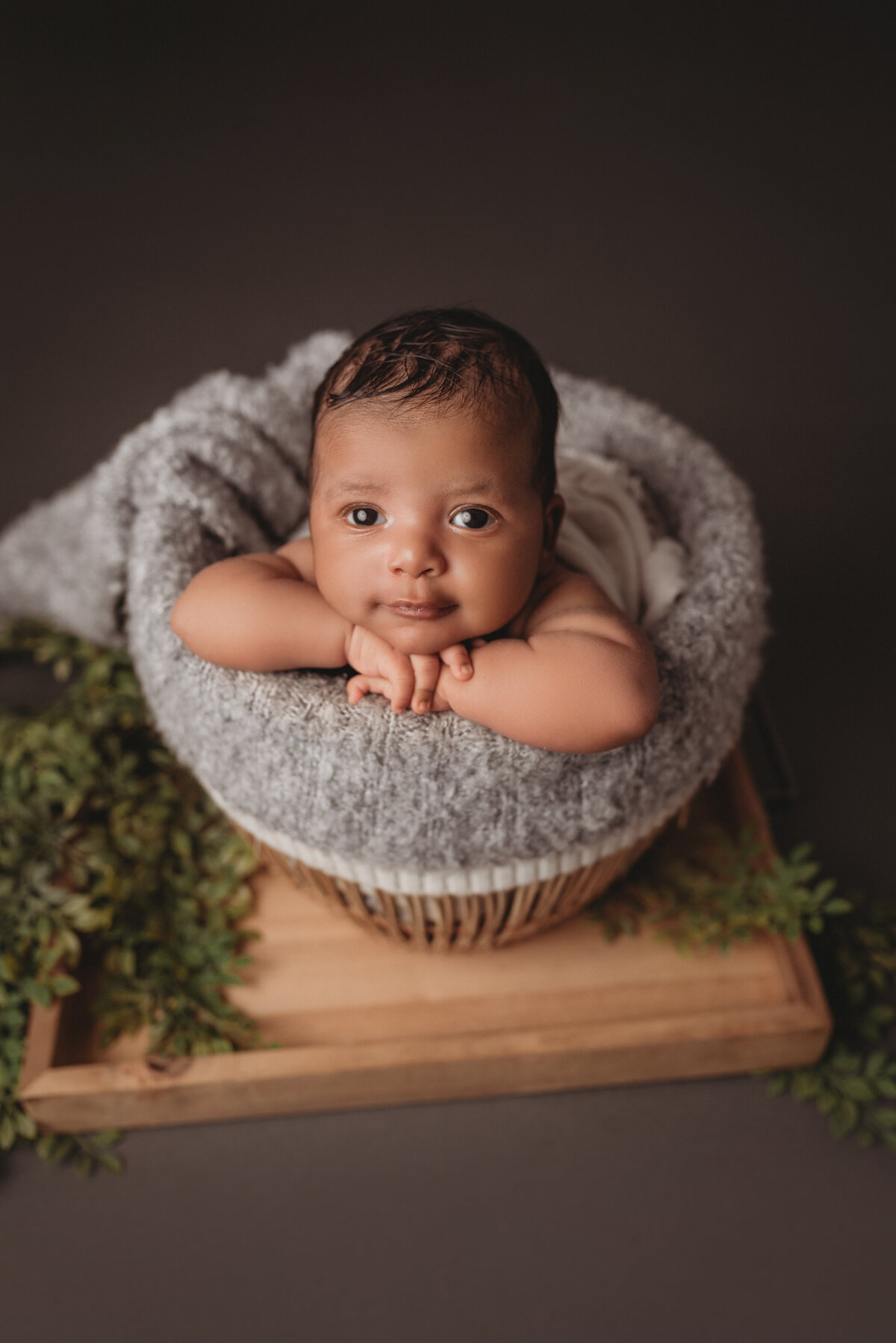 Newborn baby boy posed in wicker basket with chin on hands looking at camera with soft smile on gray backdrop