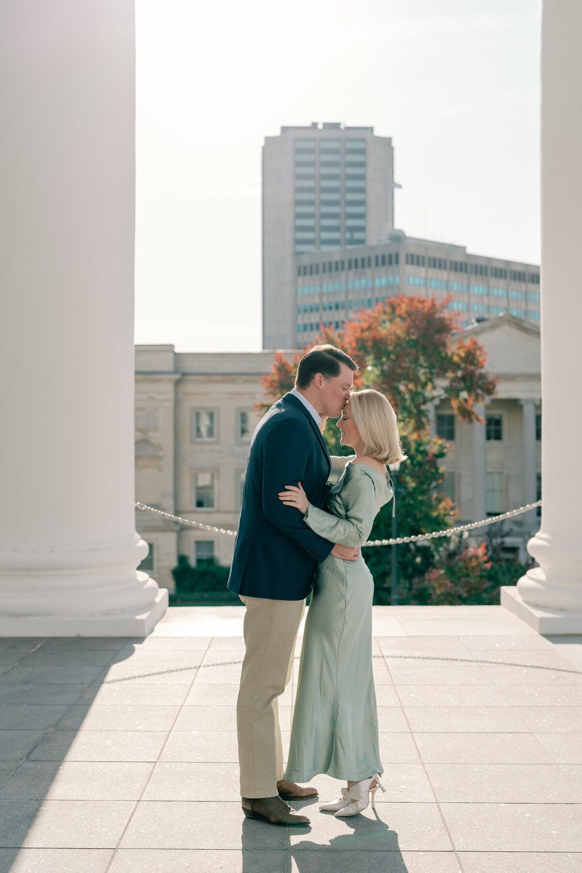 Virginia State Capitol Building Engagement Photos Richmond VA | newly engaged couple in green dress and blue suit hug in the columns of the Virginia Capitol Building in Richmond. The groom-to-be gently kisses his future wife's forehead
