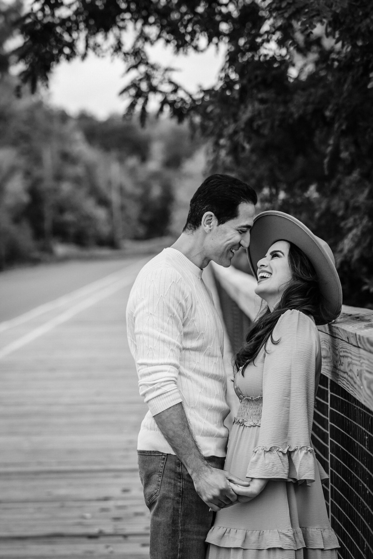 A black-and-white image of a couple standing on a wooden bridge, leaning in close with their foreheads touching, both smiling affectionately at each other.