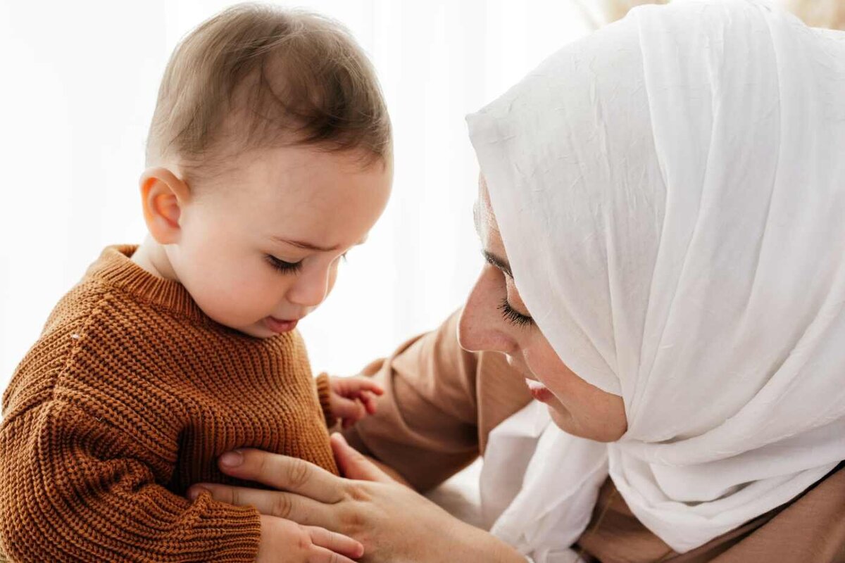 Ruua Hamdan Photography (UK): A mother wearing a white hijab interacts with her baby, who is wearing a brown sweater.