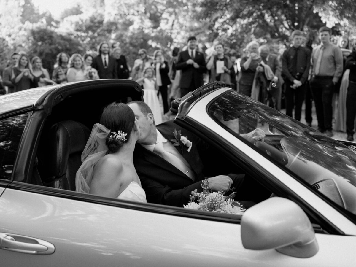 Couple's ceremony exit in a sports car with guests cheering in the background.