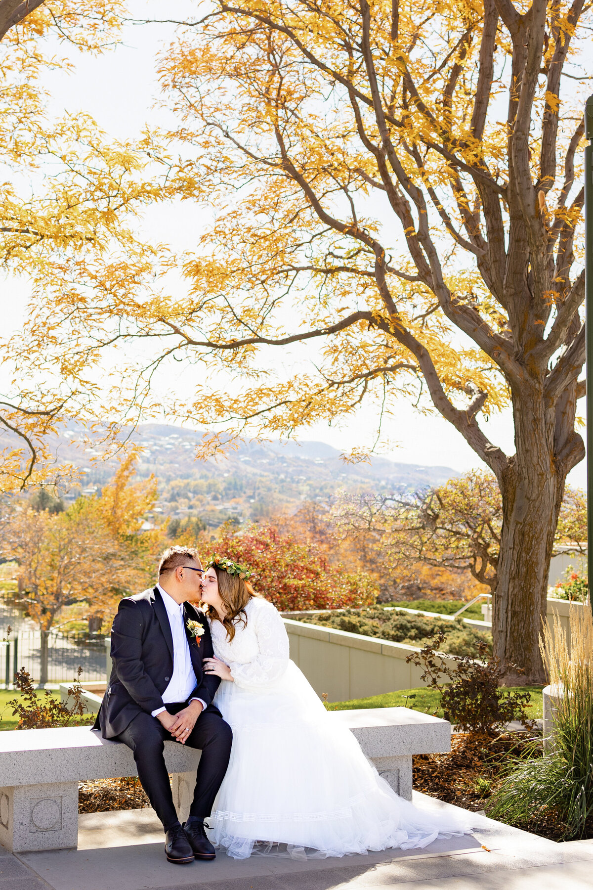 Surrounded by orange autumn leaves, the bride and groom share a kiss on a bench at the Bountiful Utah LDS Temple.