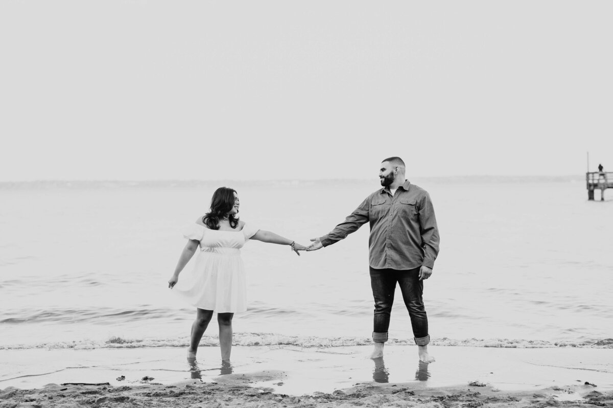 black and white engagemetn pictures with couple reaching for one another to hold hands with the water behind them