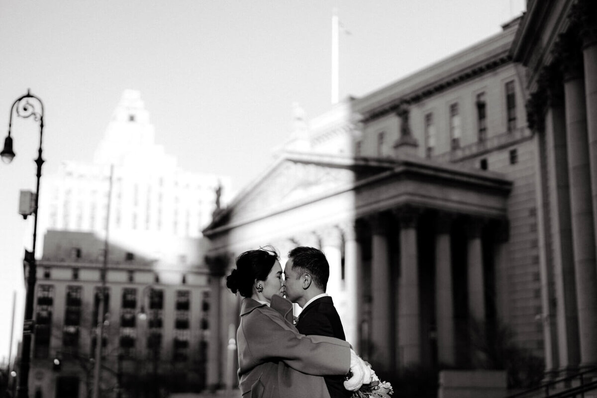 Half-body black and white photo of the bride, kissing while hugging the groom, in front of an old building with columns.