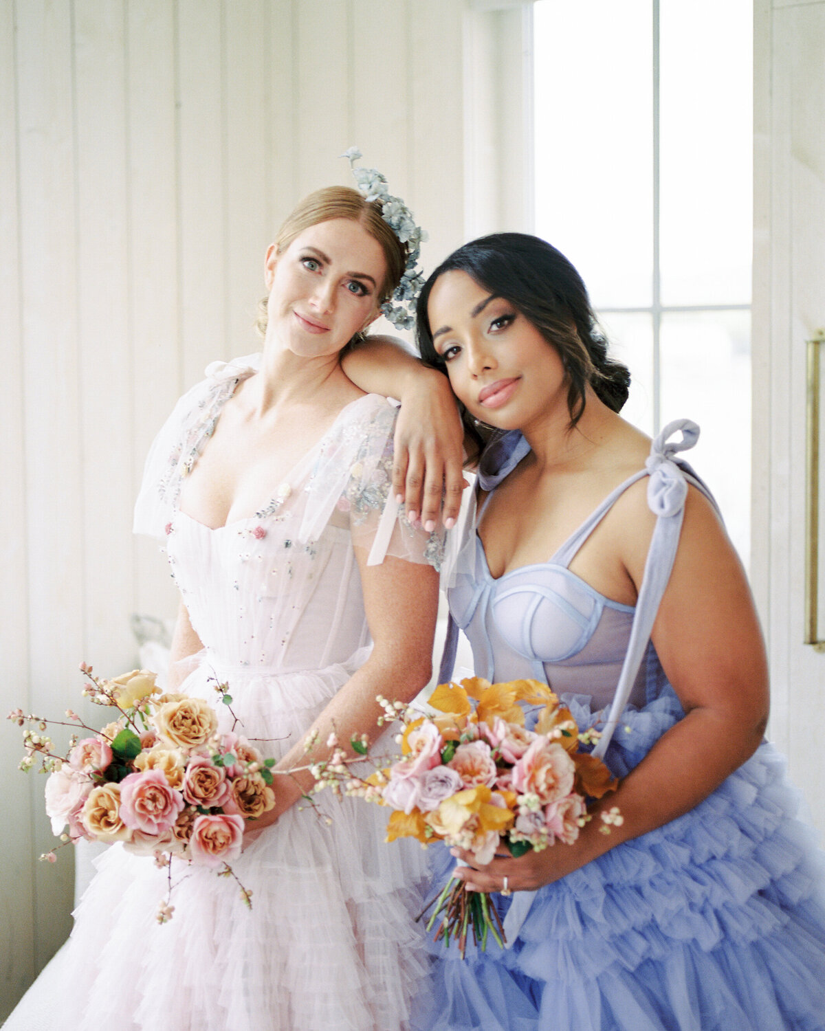 A bridesmaid wearing a lavender dress leans on the bride as they both look at the camera