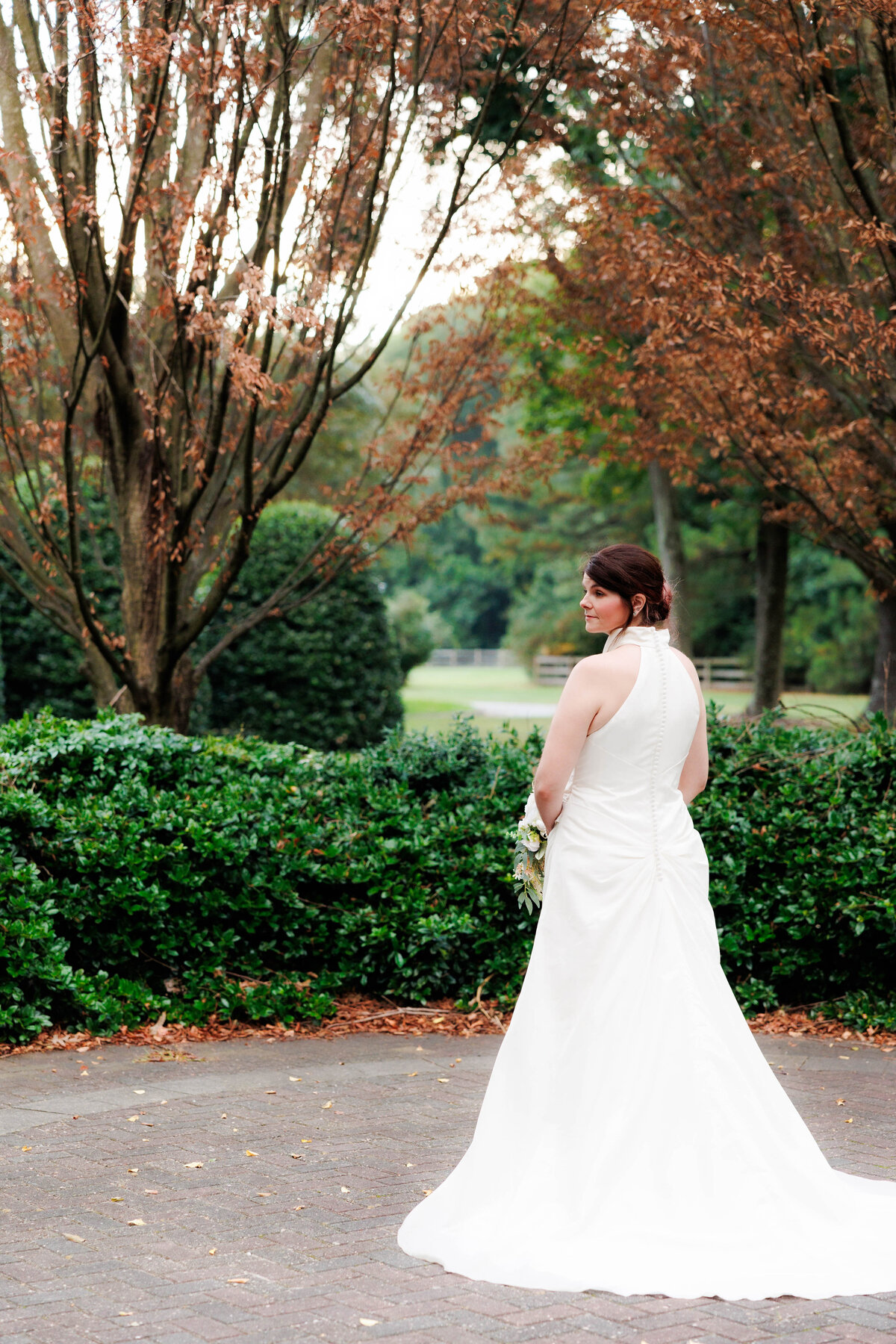 bride in the gardens of founders inn in the fall,  strapless bridal gown with open shoulders.  bride is holding bouquet and looking over shoulders but back is turned to the photographer