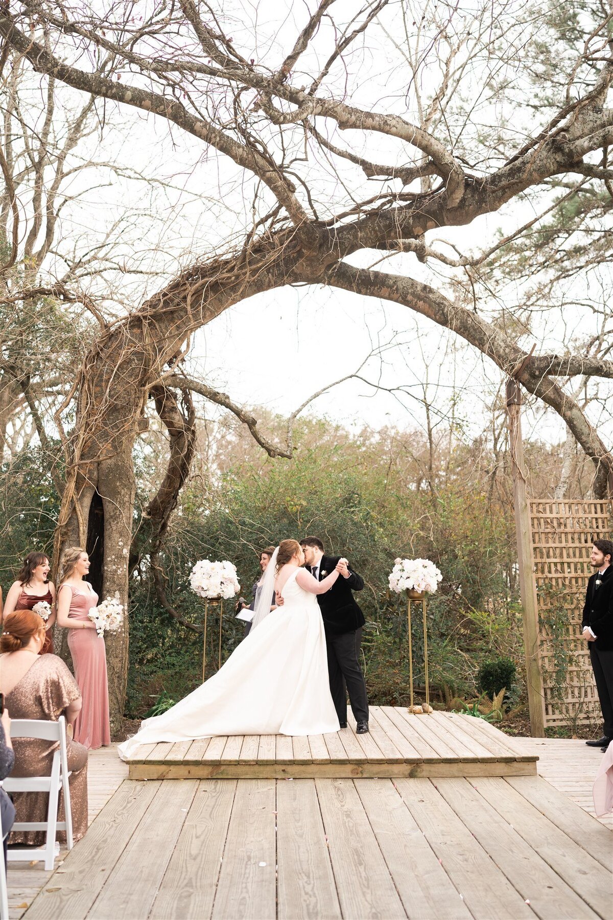 Bride and groom share their first kiss with two white floral arrangements under a large tree.