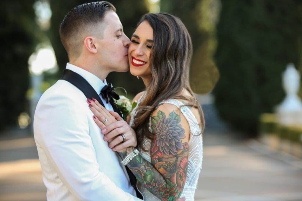 Groom kisses bride's cheek at grand island mansion with bride smiling and holding hands. Photo captured near Sacramento by philippe studio pro.