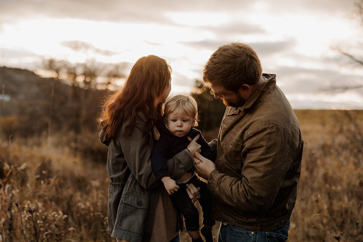 A man and woman stand outdoors in a grassy field at sunset, facing each other while tending to their young child. The woman with long hair, the man adjusting the child's jacket—captured beautifully by a New England Family Photographer. An overcast sky casts a warm glow over this tender moment.