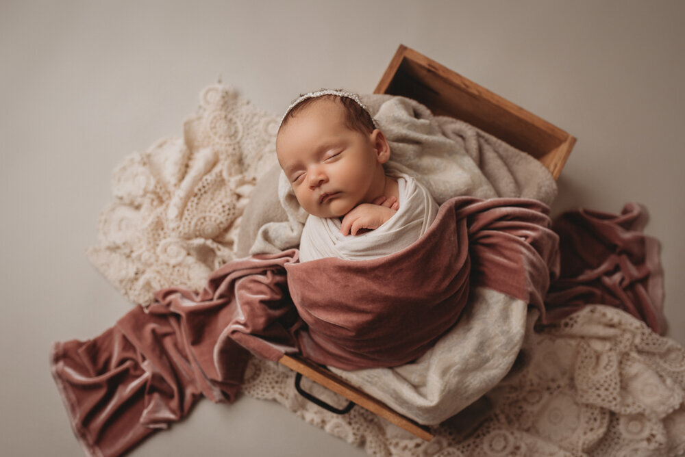 Newborn photo shoot photographed by Atlanta, Georgia newborn photographer with baby posing on neutral fabric with dainty headband on and sleeping with pink velvet fabric draped over her.