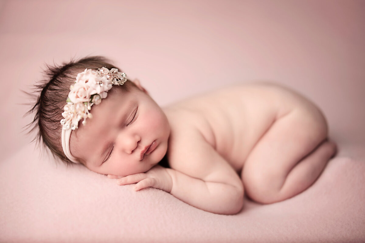 10 day old baby girl laying on tummy with legs and arms tucked under and head laying on her hand on pink fabric wearing a white flower headband
